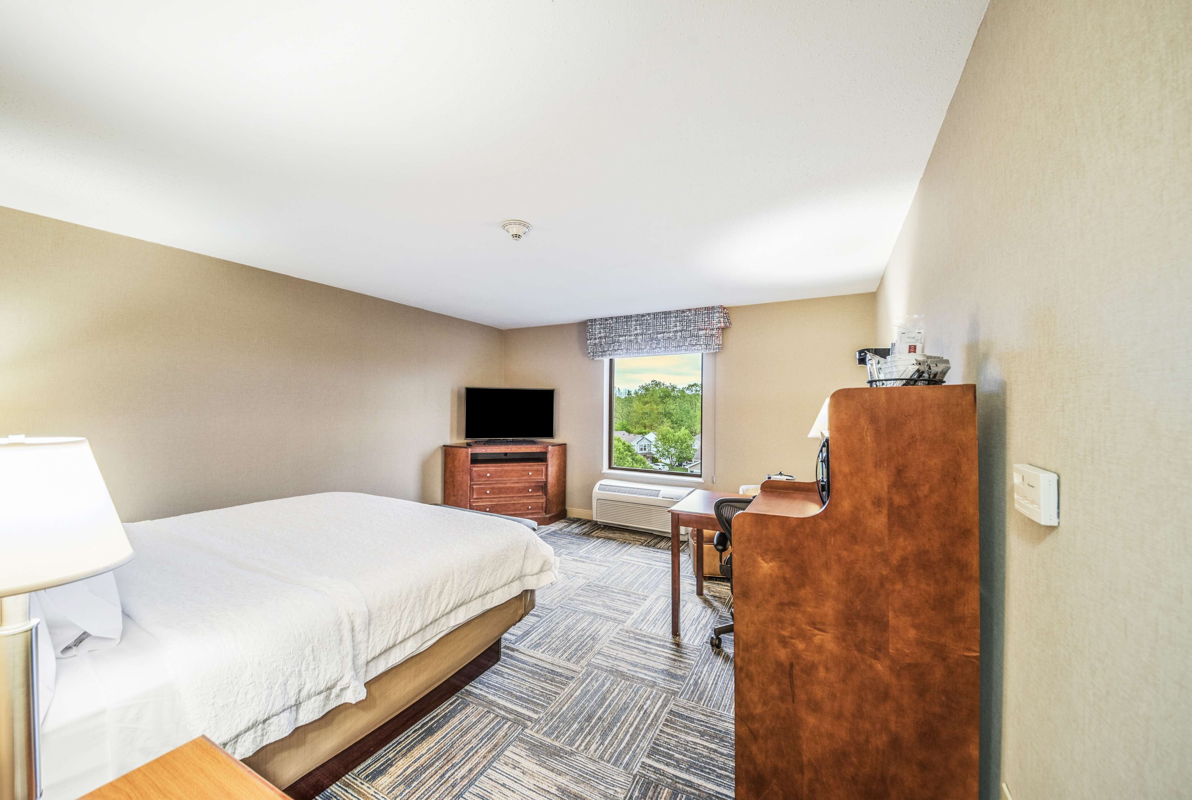 A hotel room with a bed, a desk, a television, and a window with a view of trees.