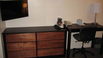 A hotel room with a wooden desk, a black office chair, a flat-screen TV mounted on the wall, a lamp, a telephone, and a coffee maker.