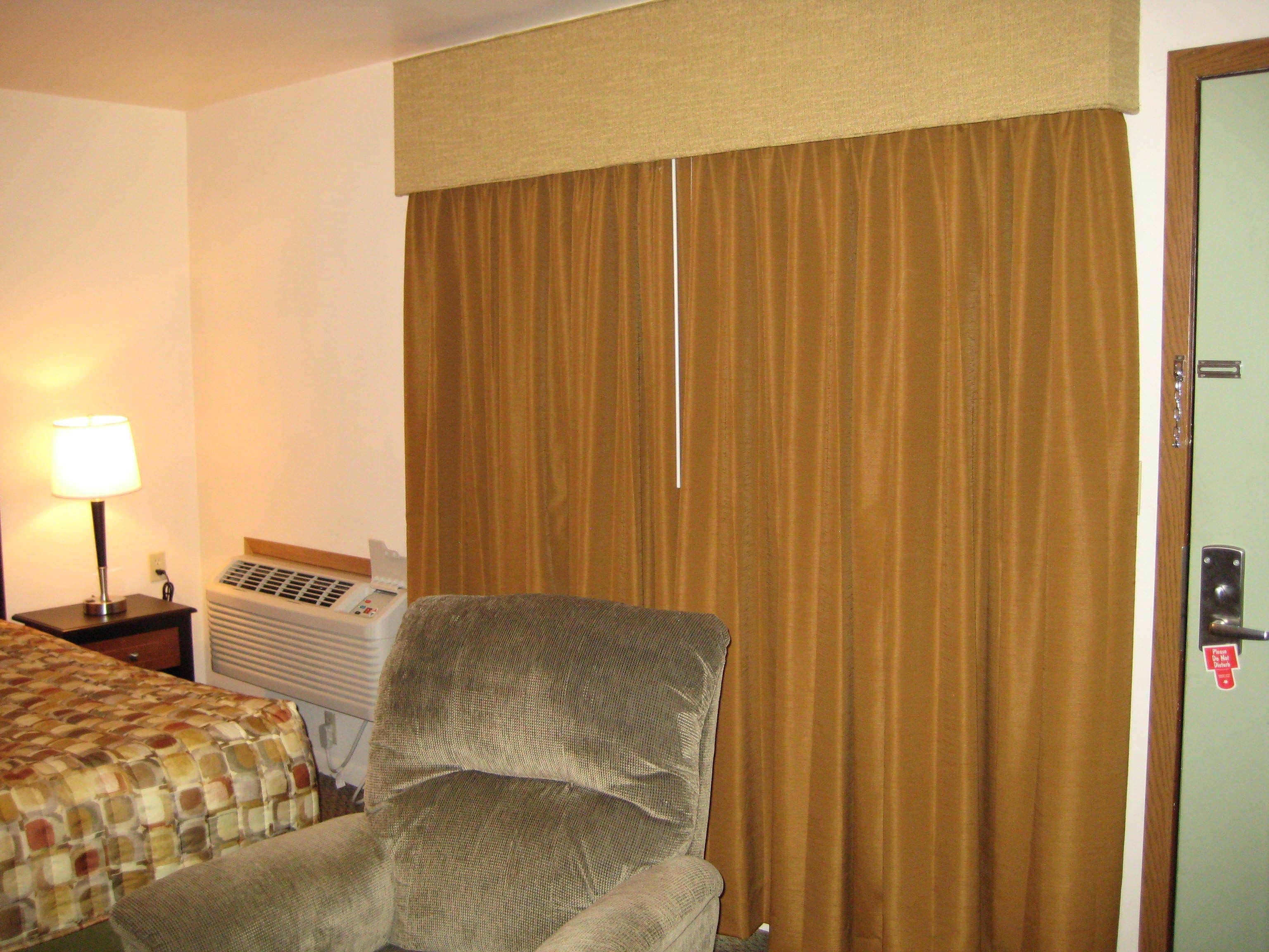 A hotel room with a bed, a chair, a lamp, and an air conditioning unit.