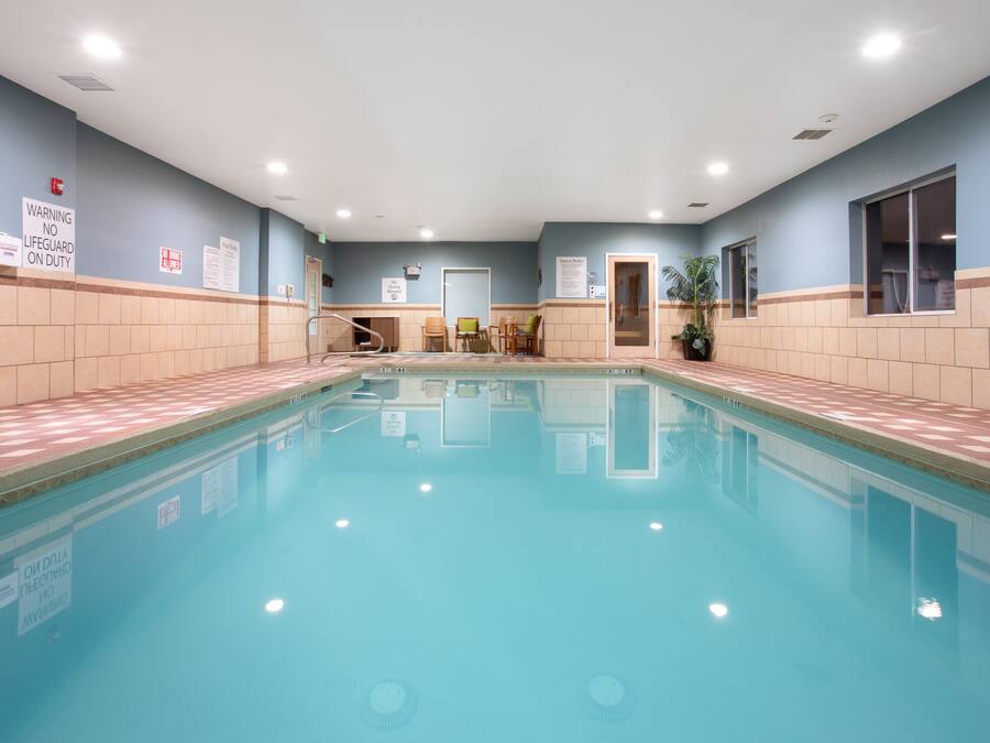 Una piscina interior con paredes de azulejos, un cartel de advertencia para socorristas y una zona de recepción con sillas y una planta.