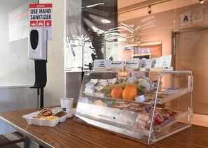 A breakfast counter with a hand sanitizer dispenser, a fruit display, and labeled food items. - Holiday Inn Express & Suites Corona by IHG (Corona)