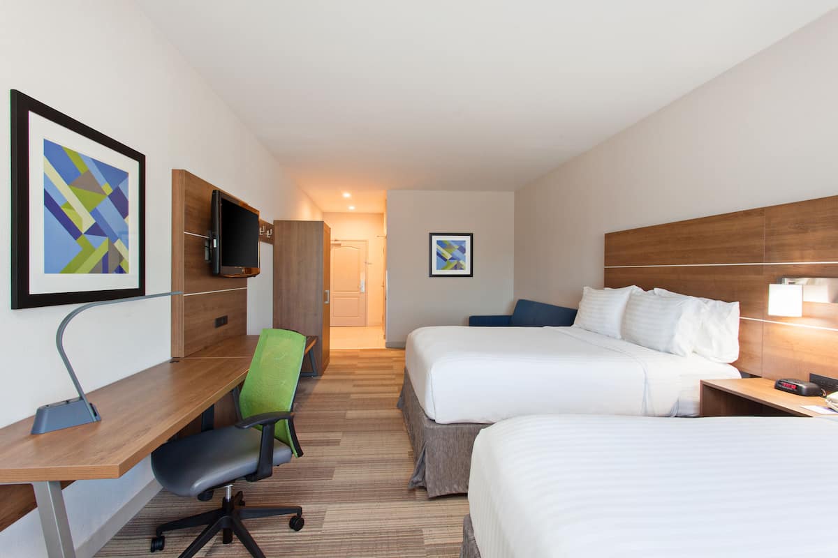 A hotel room with two beds, a desk, a chair, a TV, and artwork on the walls.