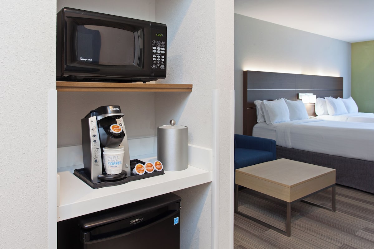 A hotel room with a microwave, coffee machine, and a small refrigerator on a white shelf. There is a bed with white bedding and a blue chair.