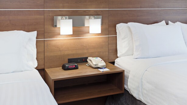 A hotel room with two beds, a wooden nightstand with a telephone and a digital clock, and wall-mounted lights. - Holiday Inn Express & Suites Corona by IHG (Corona)