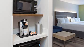 Habitación de hotel con microondas, cafetera y un refrigerador pequeño en un estante. Hay una cama con ropa blanca y una silla azul.