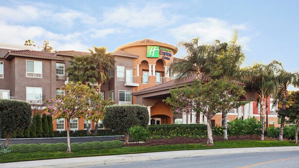 A Holiday Inn Express & Suites hotel with a brown facade, multiple windows, and a landscaped front yard with trees and shrubs. - Holiday Inn Express & Suites Corona by IHG (Corona)