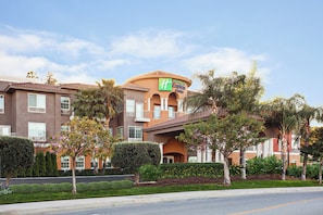 A Holiday Inn Express & Suites hotel with a brown facade, multiple windows, and a landscaped front yard with trees and shrubs. - Holiday Inn Express & Suites Corona by IHG (Corona)
