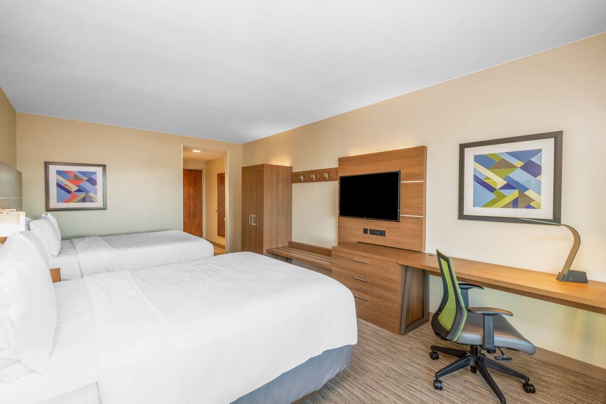 A hotel room with two beds, a desk with a chair, a flat-screen TV, and artwork on the walls.