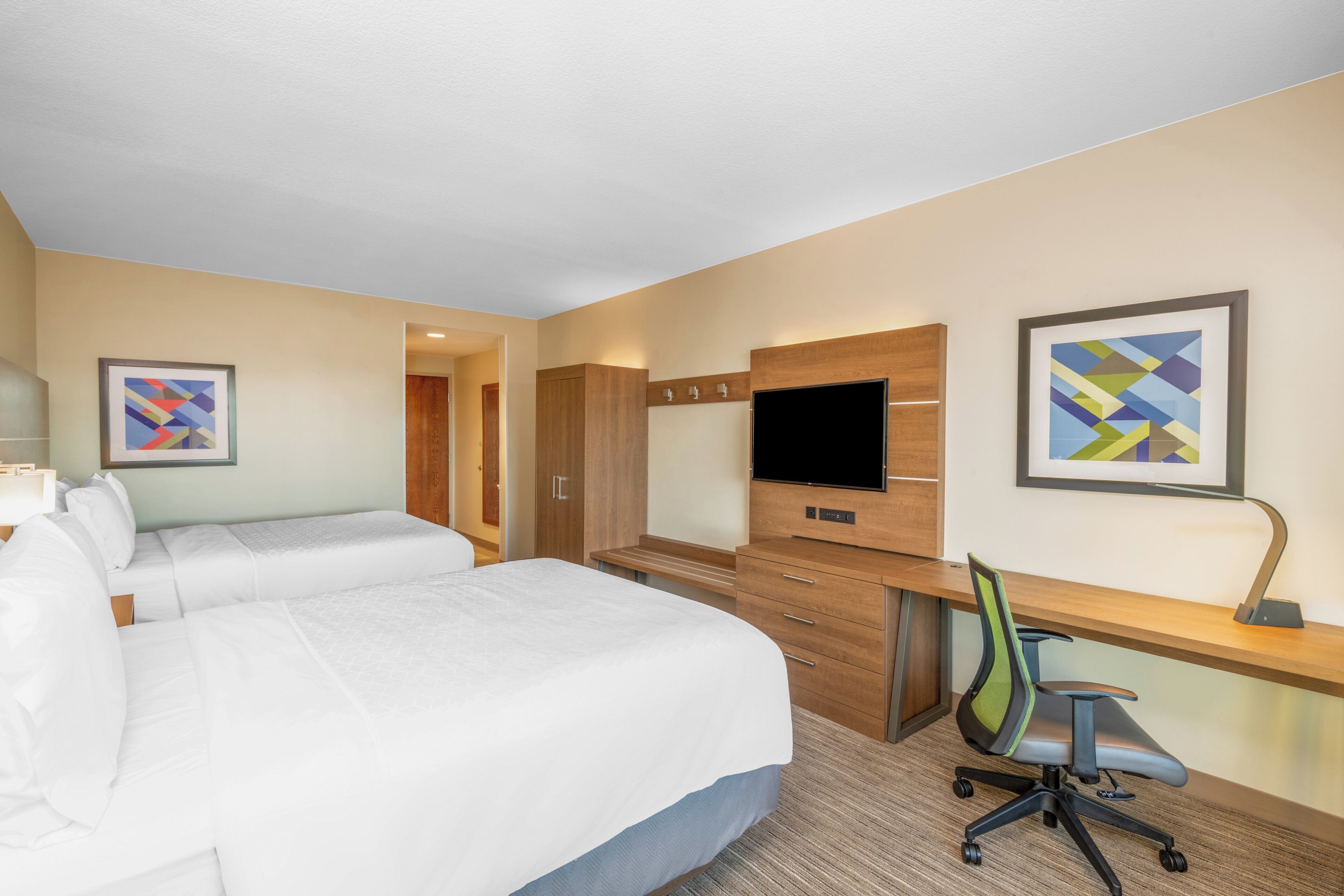 Habitación de hotel con dos camas, un escritorio con silla, televisor de pantalla plana y obras de arte en las paredes.