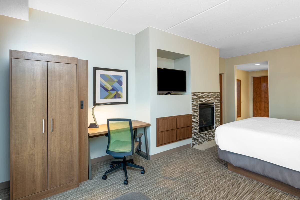 A hotel room with a bed, a desk with a chair, a television, a wardrobe, and a fireplace.