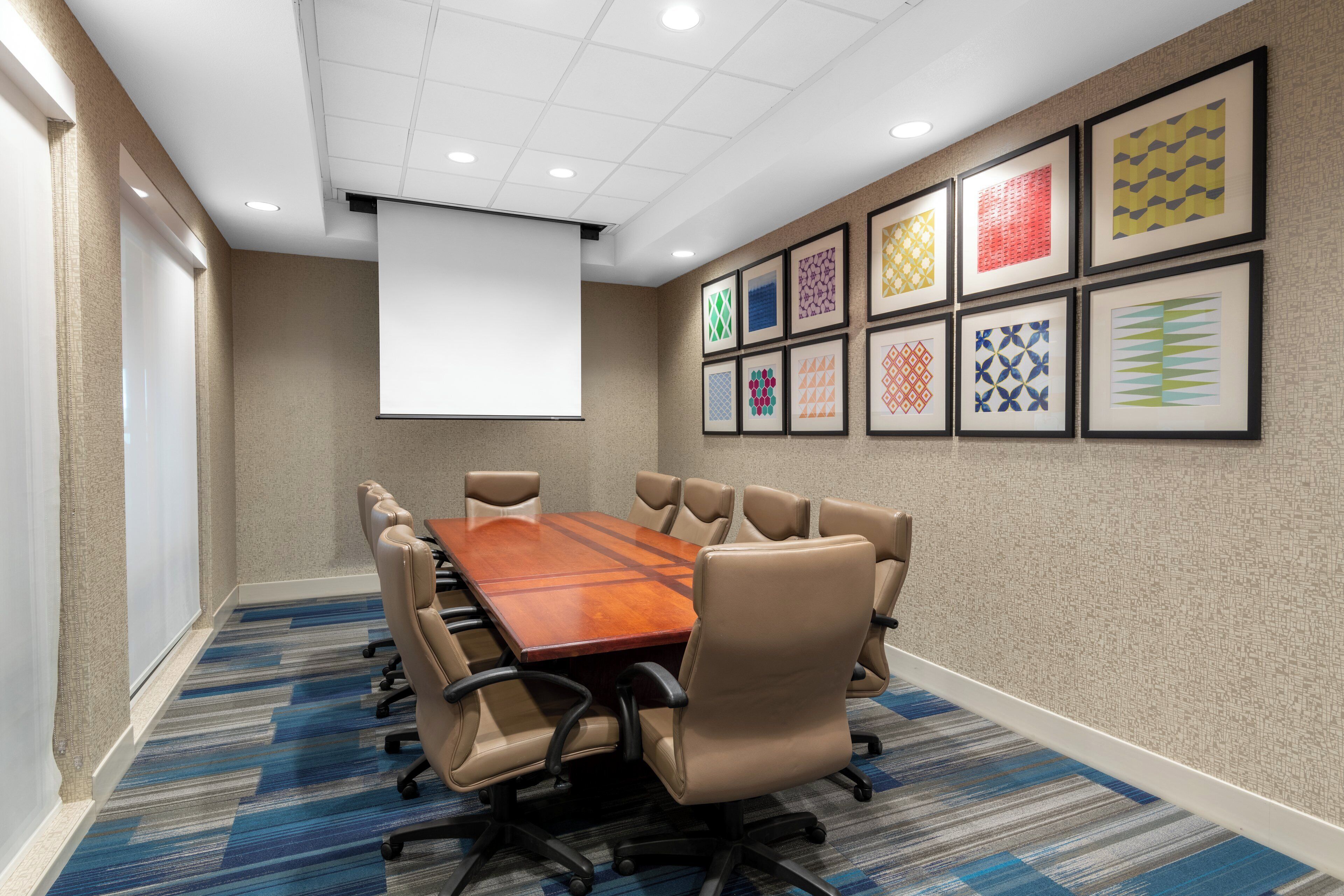 A conference room with a long table, multiple chairs, a projector screen, and patterned wall art.