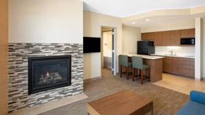 A modern living room with a stone fireplace, a TV mounted on the wall, and a kitchen area with wooden cabinets and a microwave. - Holiday Inn Express & Suites St. George North - Zion by IHG (Washington)