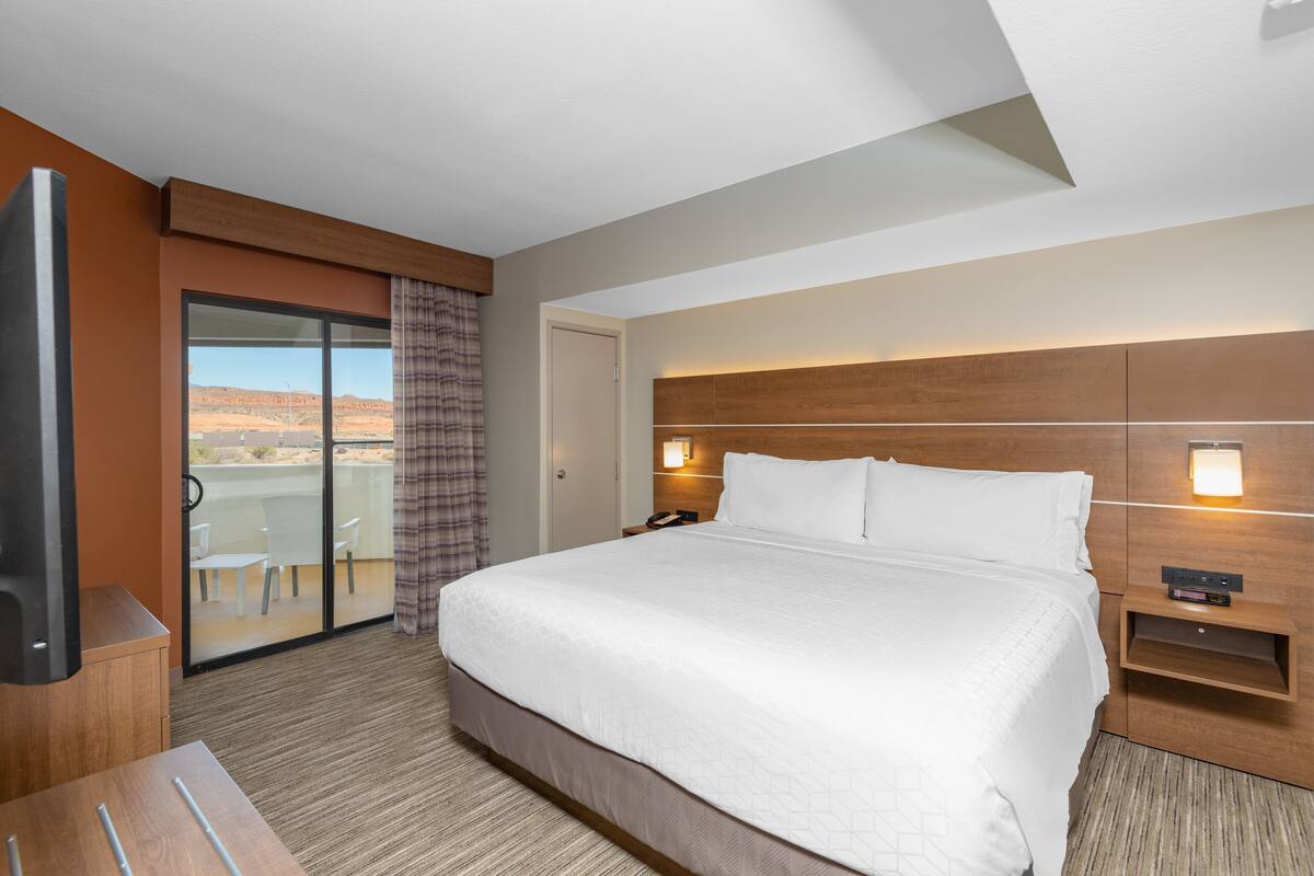 A hotel room with a large bed, a TV, a desk, and a balcony with a view of the desert.