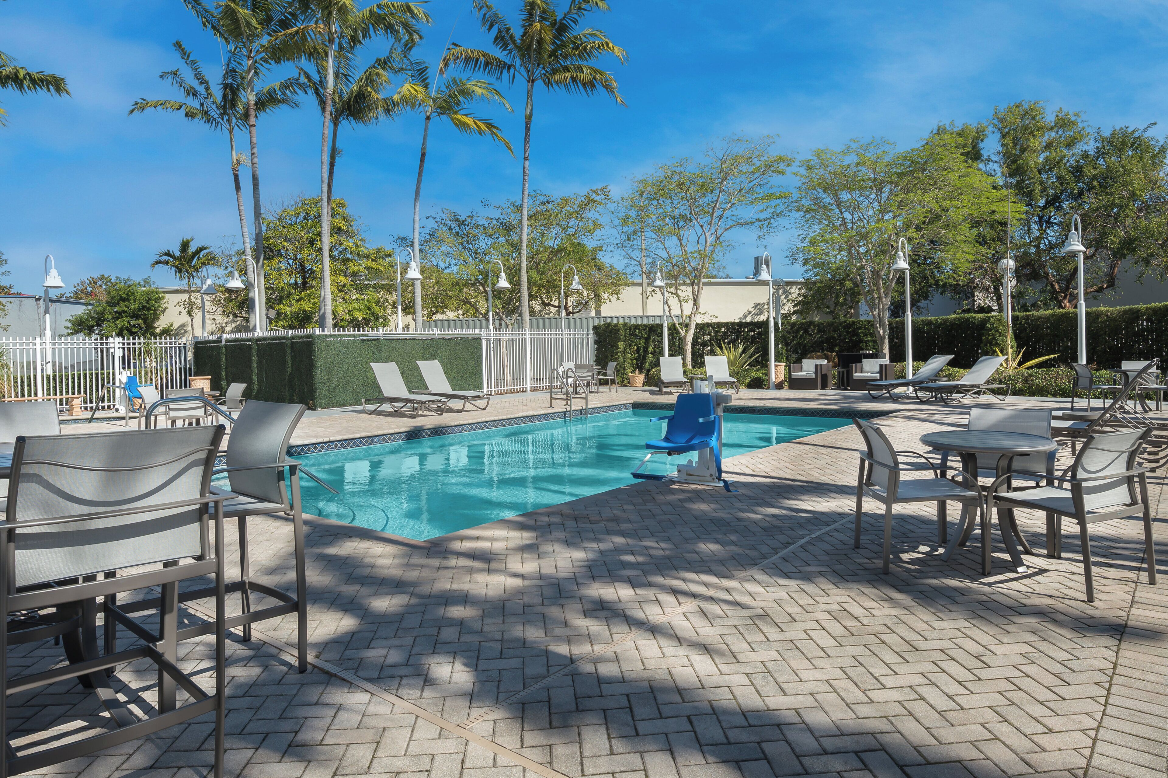 A pool area with lounge chairs and tables, surrounded by a fence and palm trees.