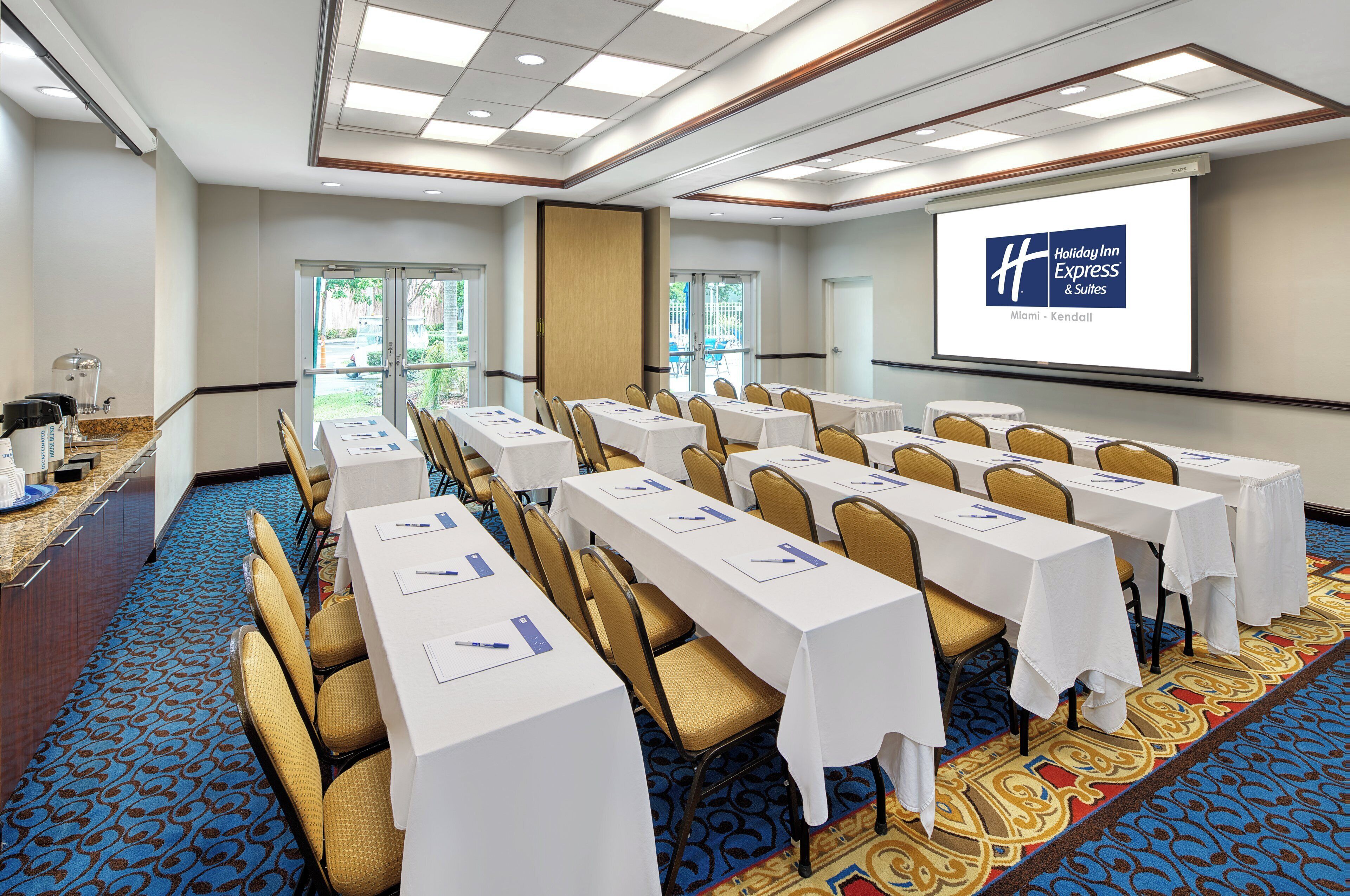 A conference room with rows of tables and chairs, a projector screen displaying a hotel name, and a water dispenser on the left.
