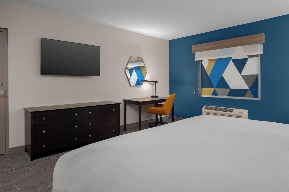 A hotel room with a bed, a desk with a chair, a dresser, a flat-screen TV, a mirror, a window with a geometric curtain, and an air conditioning unit.