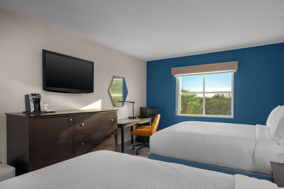 Quarto de hotel com duas camas, uma escrivaninha com computador, televisão e janela com vista.
