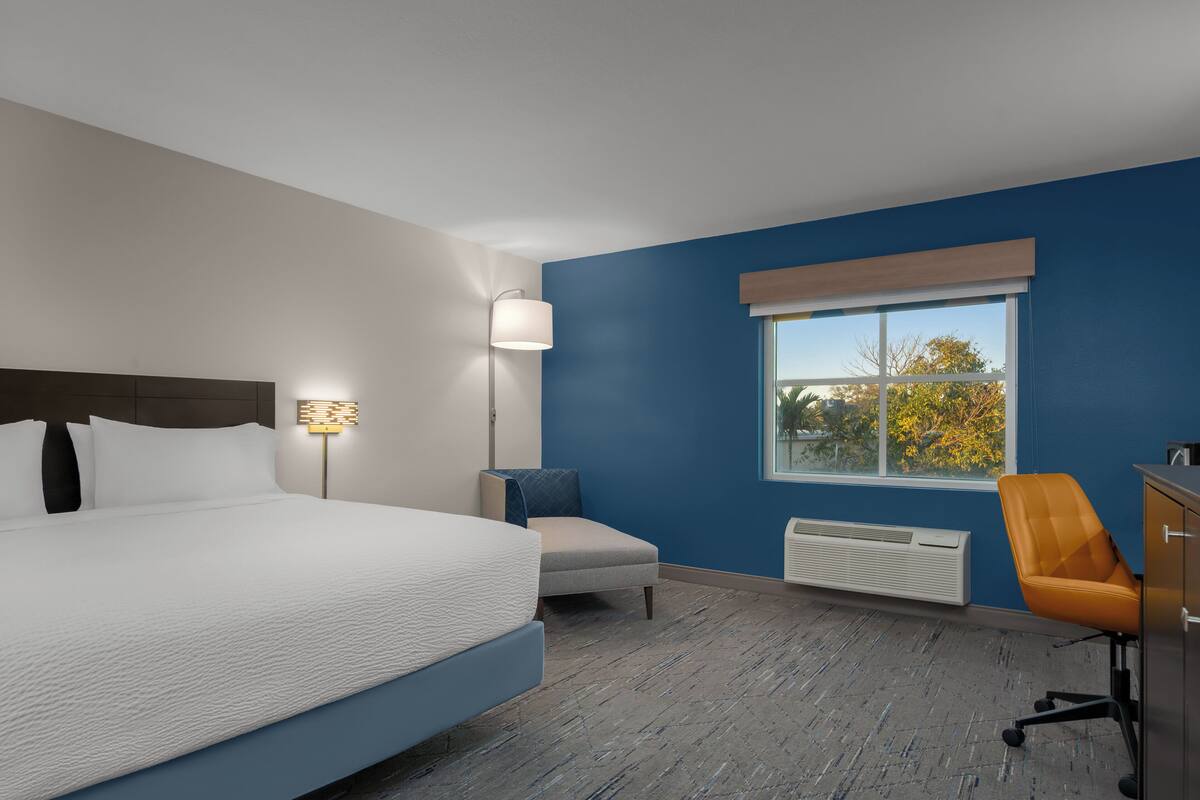 A modern hotel room with a large bed, a desk, a chair, a lamp, and a window with a view.