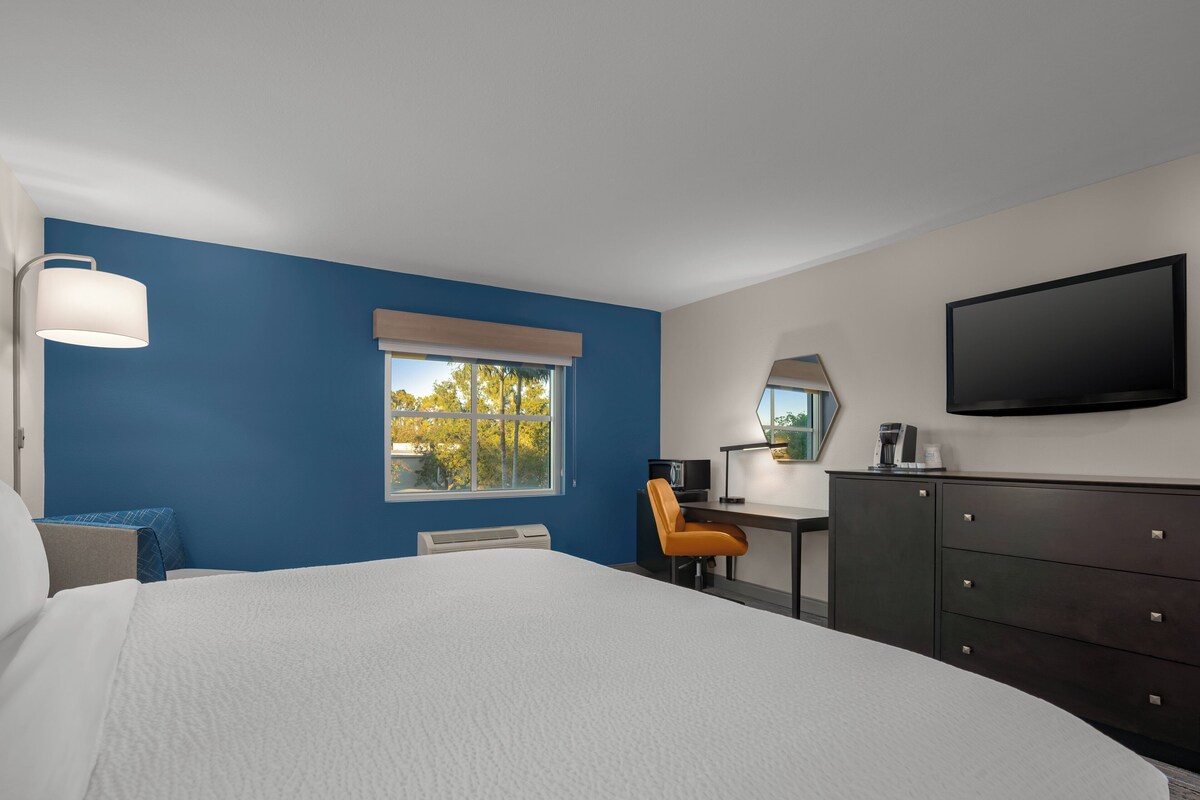 Quarto de hotel com cama, escrivaninha, cadeira, cômoda, televisão e janela com vista para árvores.