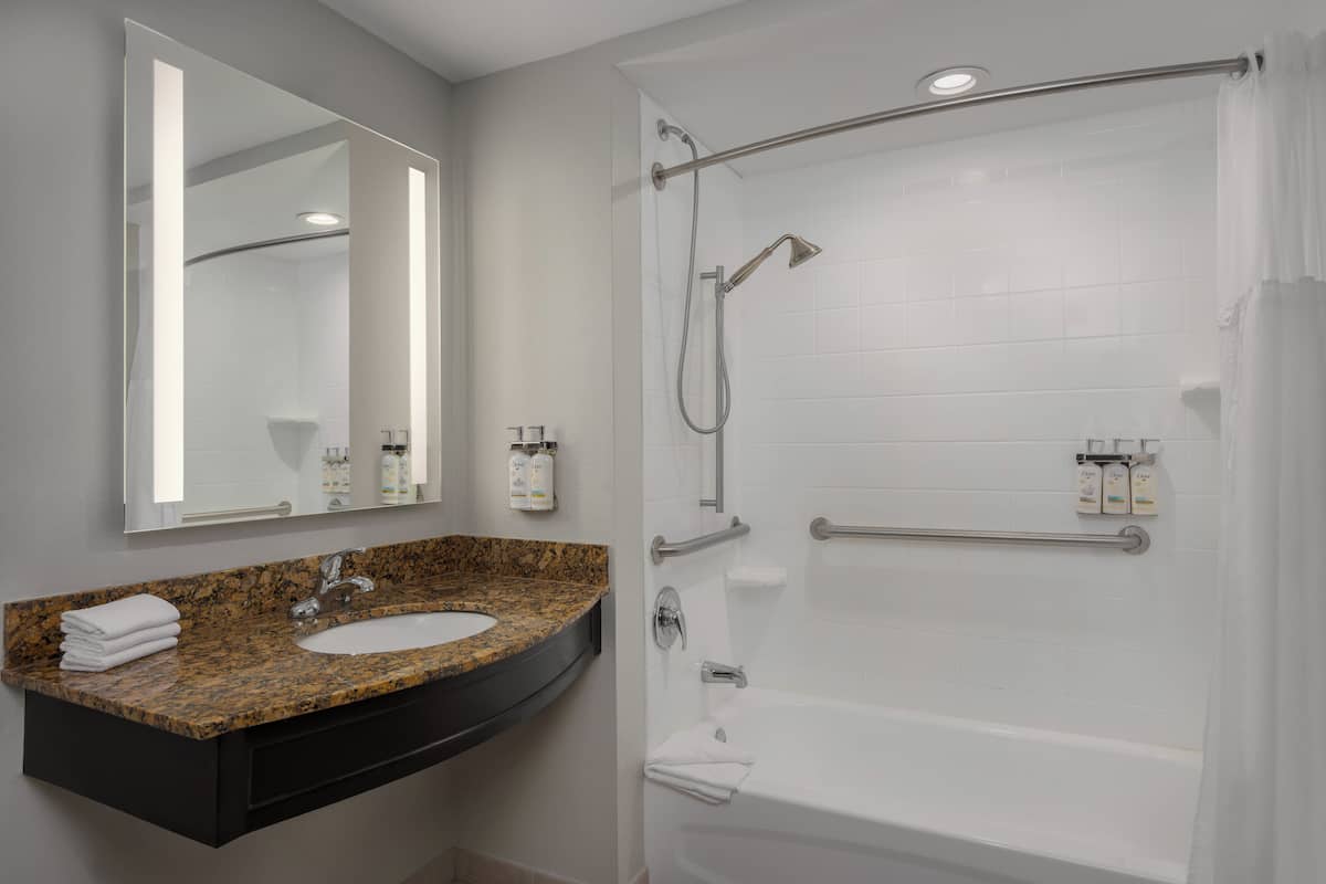A bathroom with a granite countertop sink, a mirror, a shower with grab bars, and a white bathtub.