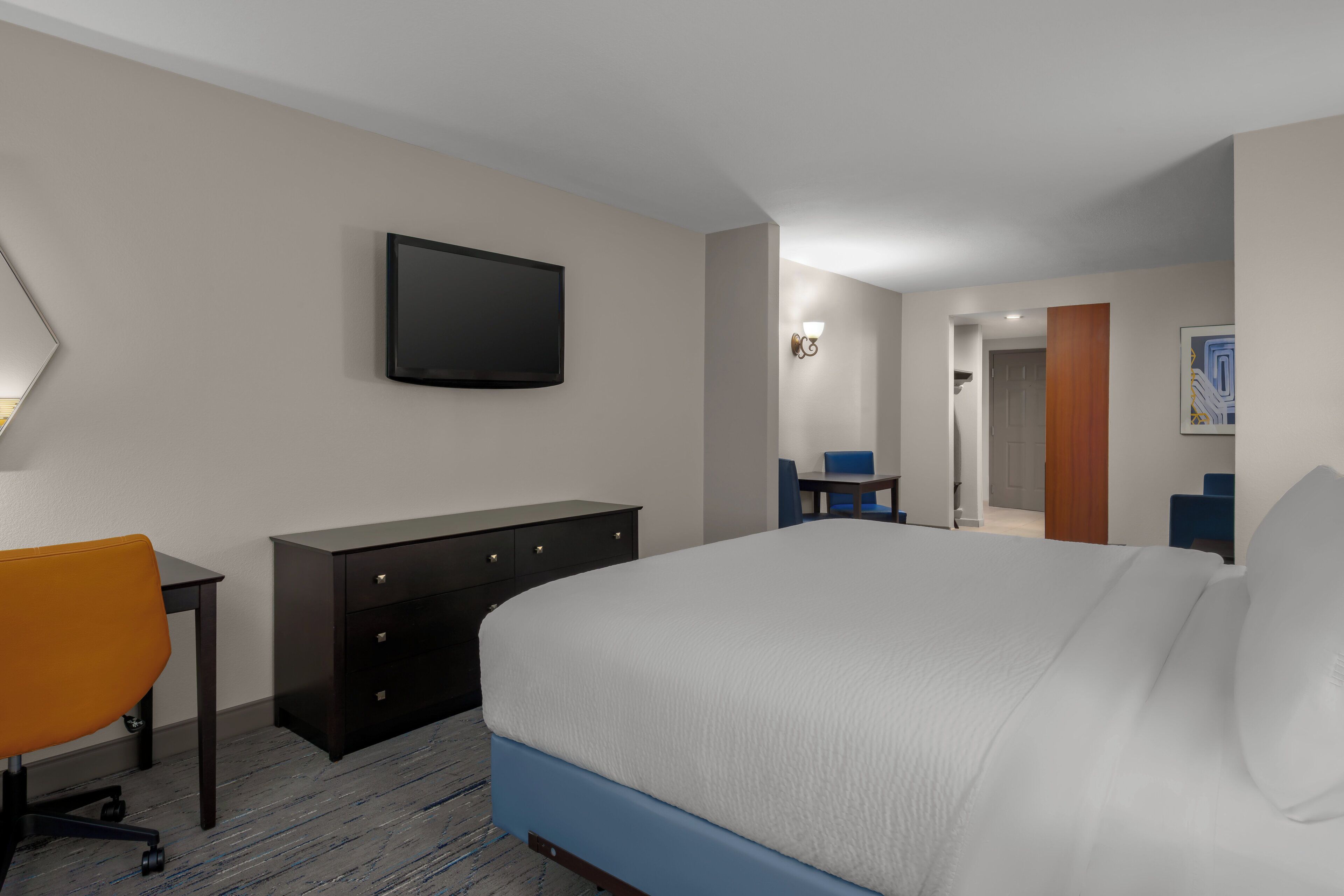 A hotel room with a bed, a desk, a chair, a dresser, a television, and a door leading to another room.
