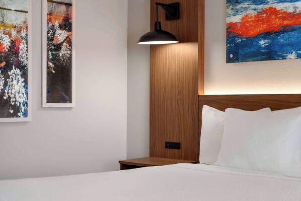 Una habitación de hotel con cabecera de madera, lámpara de noche, cama blanca y dos cuadros enmarcados en las paredes.