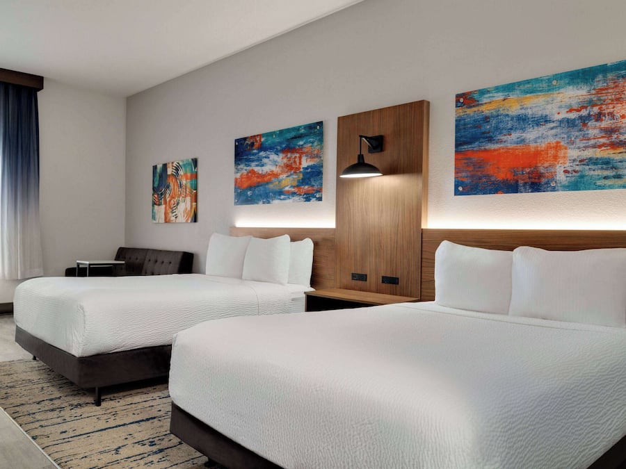 Quarto de hotel com duas camas, cabeceira de madeira, luminária na parede e quadros abstratos.