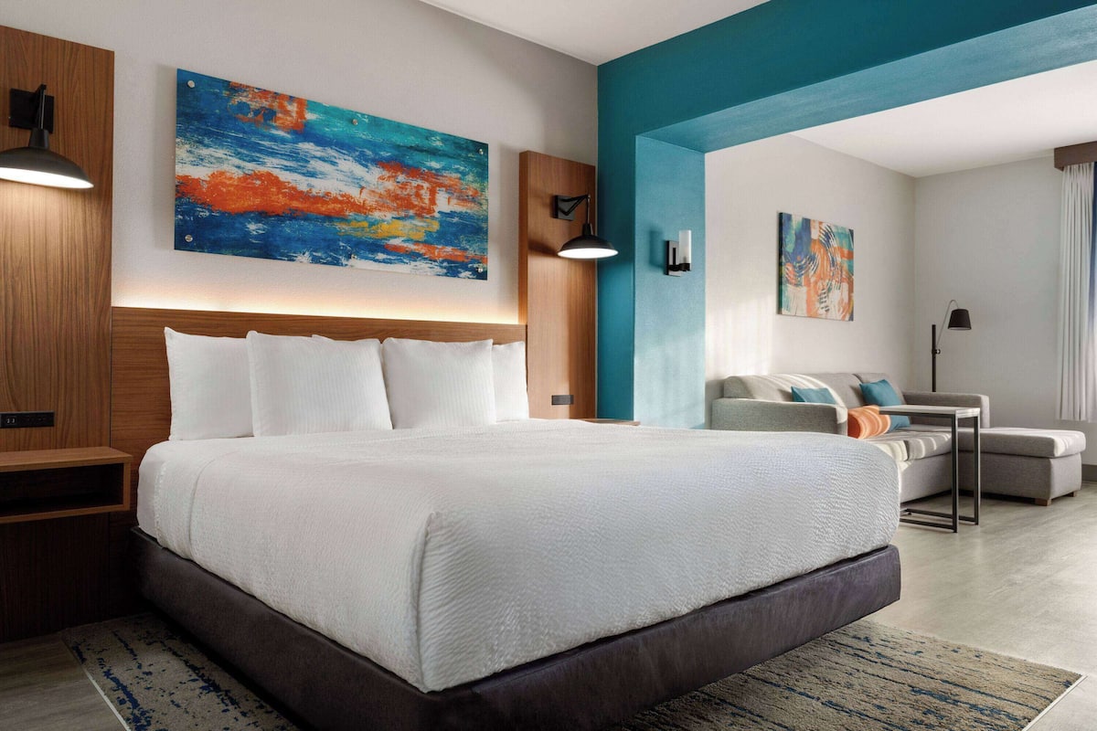 Een moderne hotelkamer met een groot bed, een bureau en een bank. Twee schilderijen hangen aan de muur en er staat een lamp op het bureau.