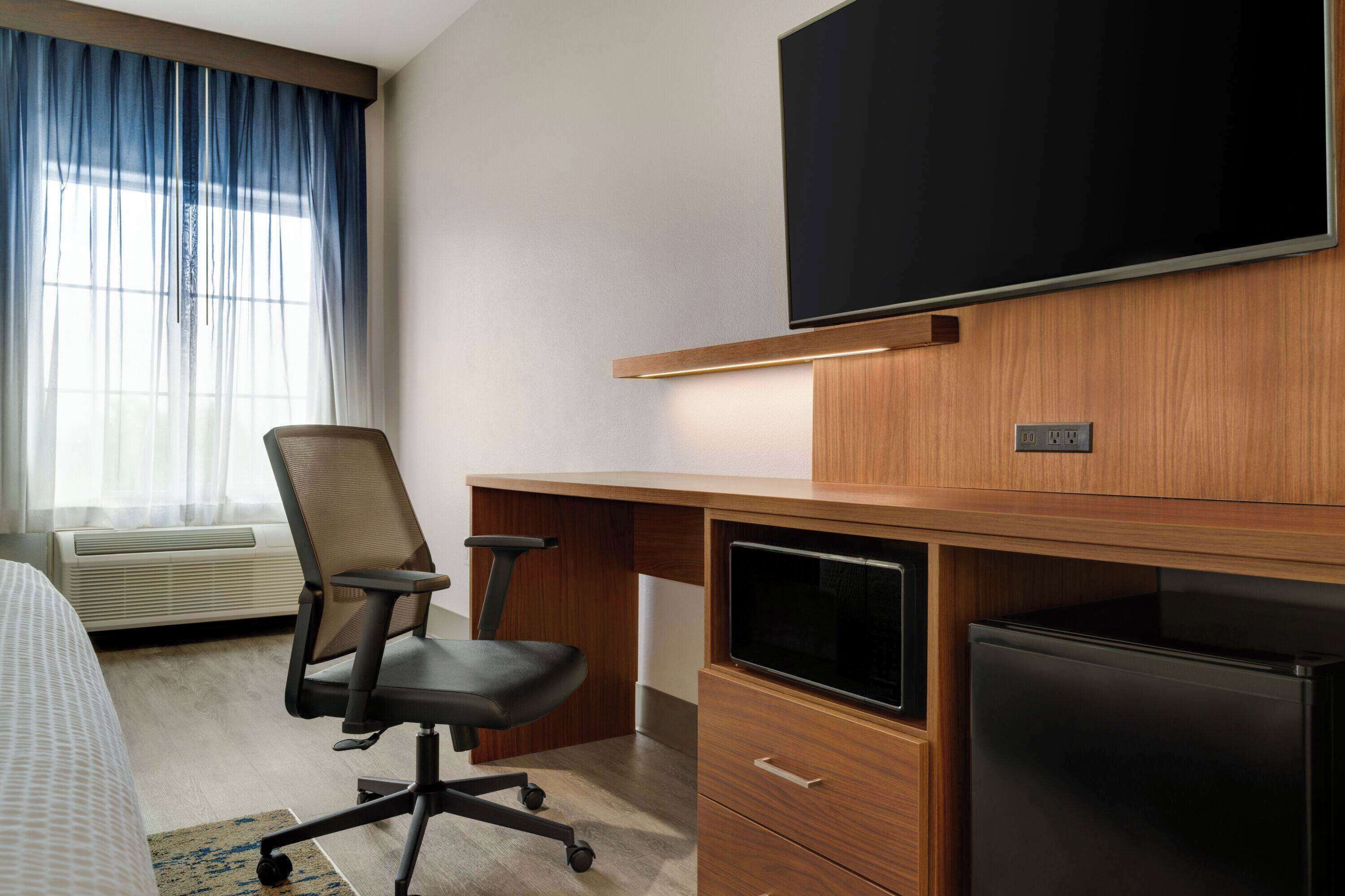 A hotel room with a bed, a desk with a computer, a TV, and a refrigerator.