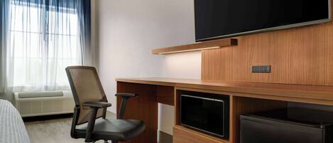 A hotel room with a bed, a desk with a computer, a TV, and a refrigerator.