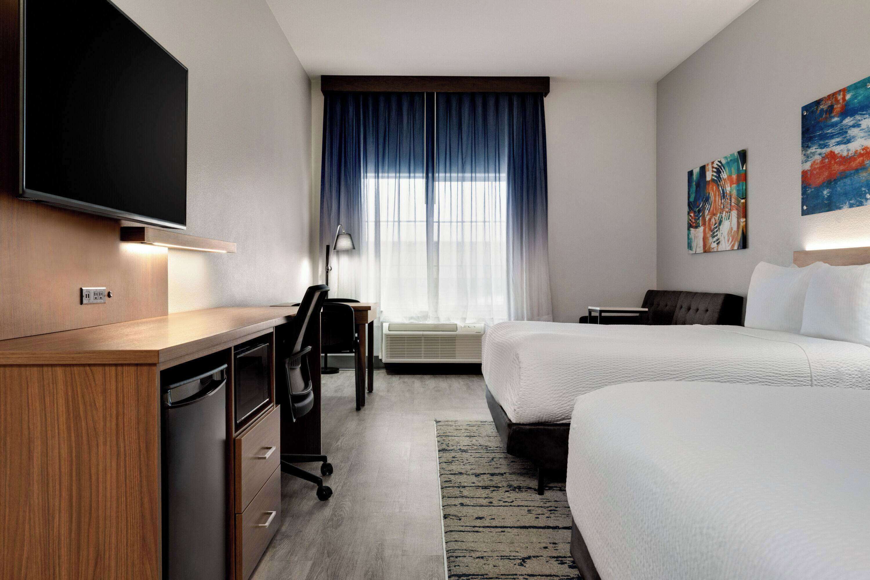Habitación de hotel con televisión de pantalla plana, un escritorio con silla, dos camas, un ventanal grande con cortinas y obras de arte en la pared.