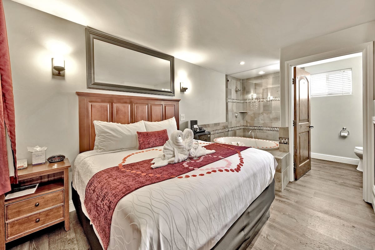 A hotel room with a large bed, a wooden nightstand, a bathroom with a shower, and a mirror above the bed.