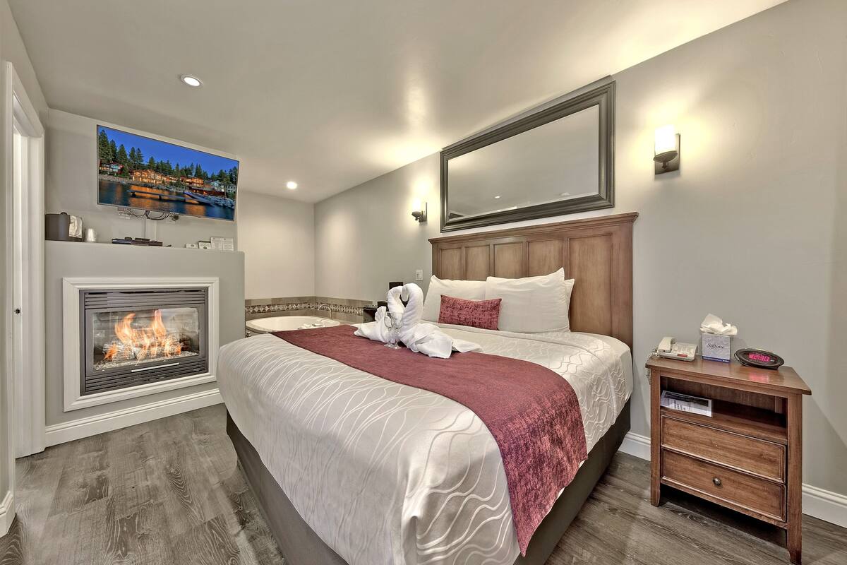 A hotel room with a large bed, a fireplace, a nightstand, and a wall-mounted TV.