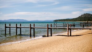 Une plage avec des jetées en bois s’avançant dans des eaux calmes, une rive sablonneuse et une colline boisée en arrière-plan.