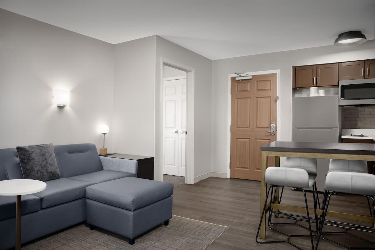 A modern living room with a sectional sofa, a small dining table with bar stools, a kitchen area with a refrigerator and microwave, and a door leading to another room.