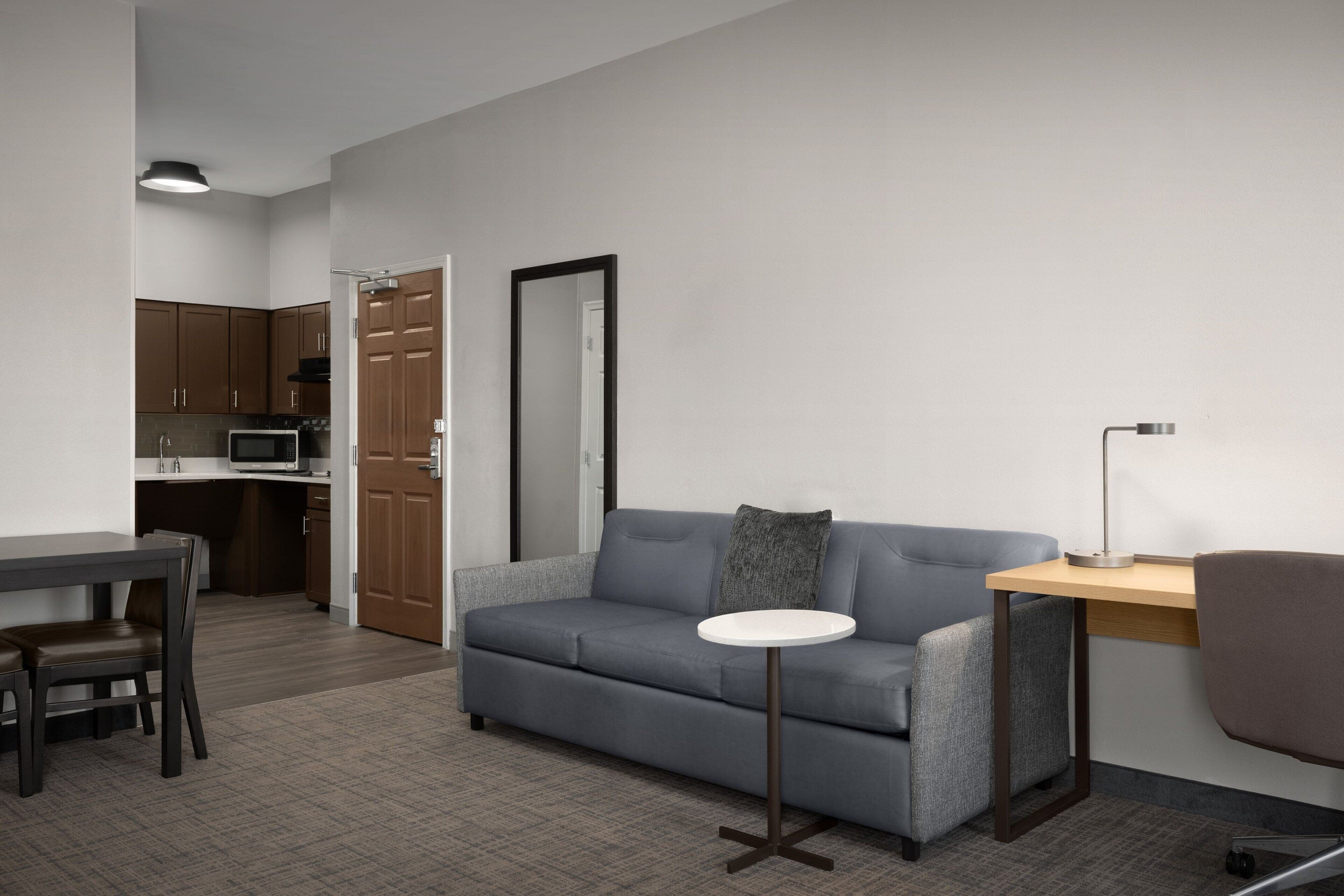 Ein modernes Wohnzimmer mit einem grauen Sofa, einem kleinen runden Tisch, einem Schreibtisch mit Lampe und einer Küchenzeile im Hintergrund.