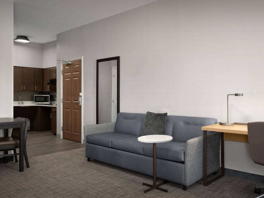 Ein modernes Wohnzimmer mit einem grauen Sofa, einem kleinen runden Tisch, einem Schreibtisch mit Lampe und einer Küchenzeile im Hintergrund.