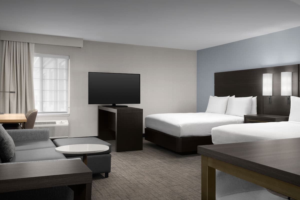 A modern hotel room with a flat-screen TV, two beds, a sofa, a small table, and a desk.