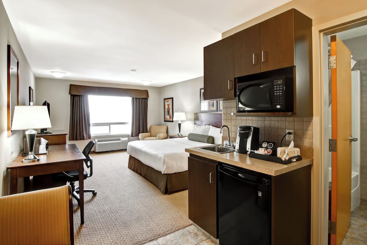 A hotel room with a bed, desk, chair, kitchenette, and a bathroom visible through an open door.
