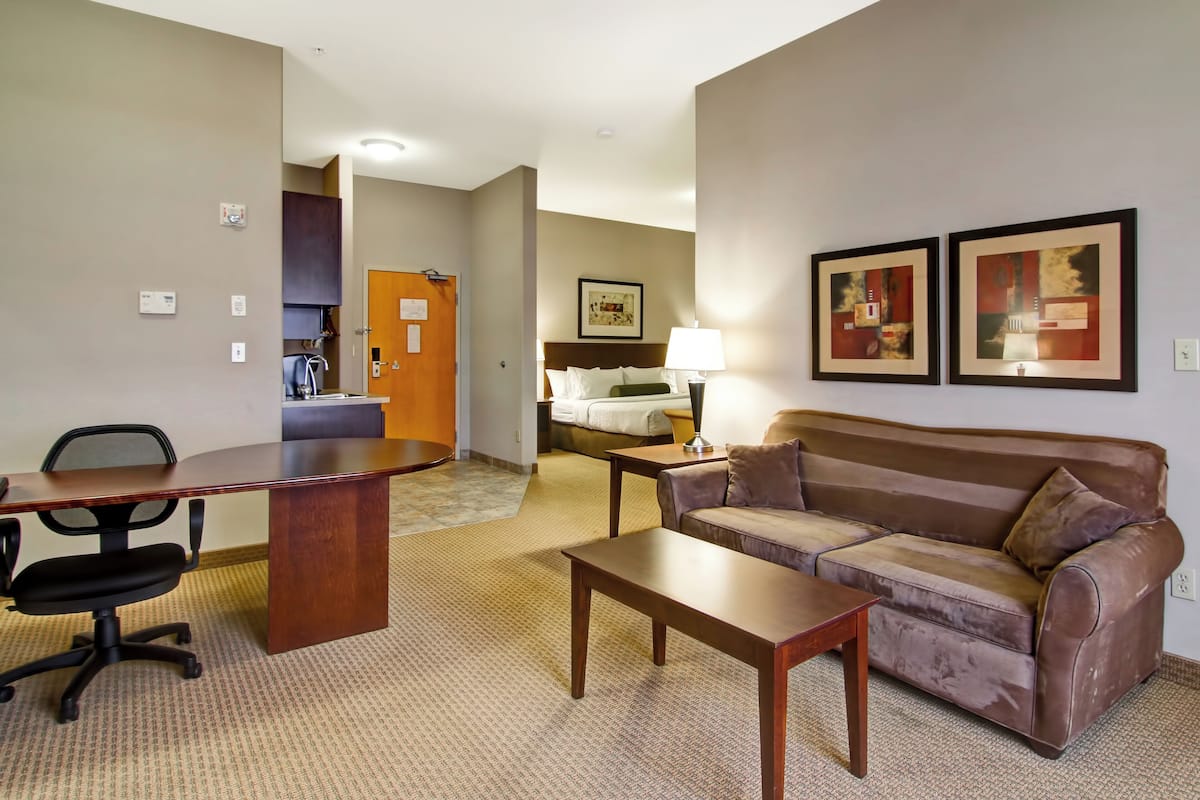 A hotel room with a wooden desk, a brown leather sofa, a bed with white linens, a small table with a lamp, and two framed pictures on the wall.
