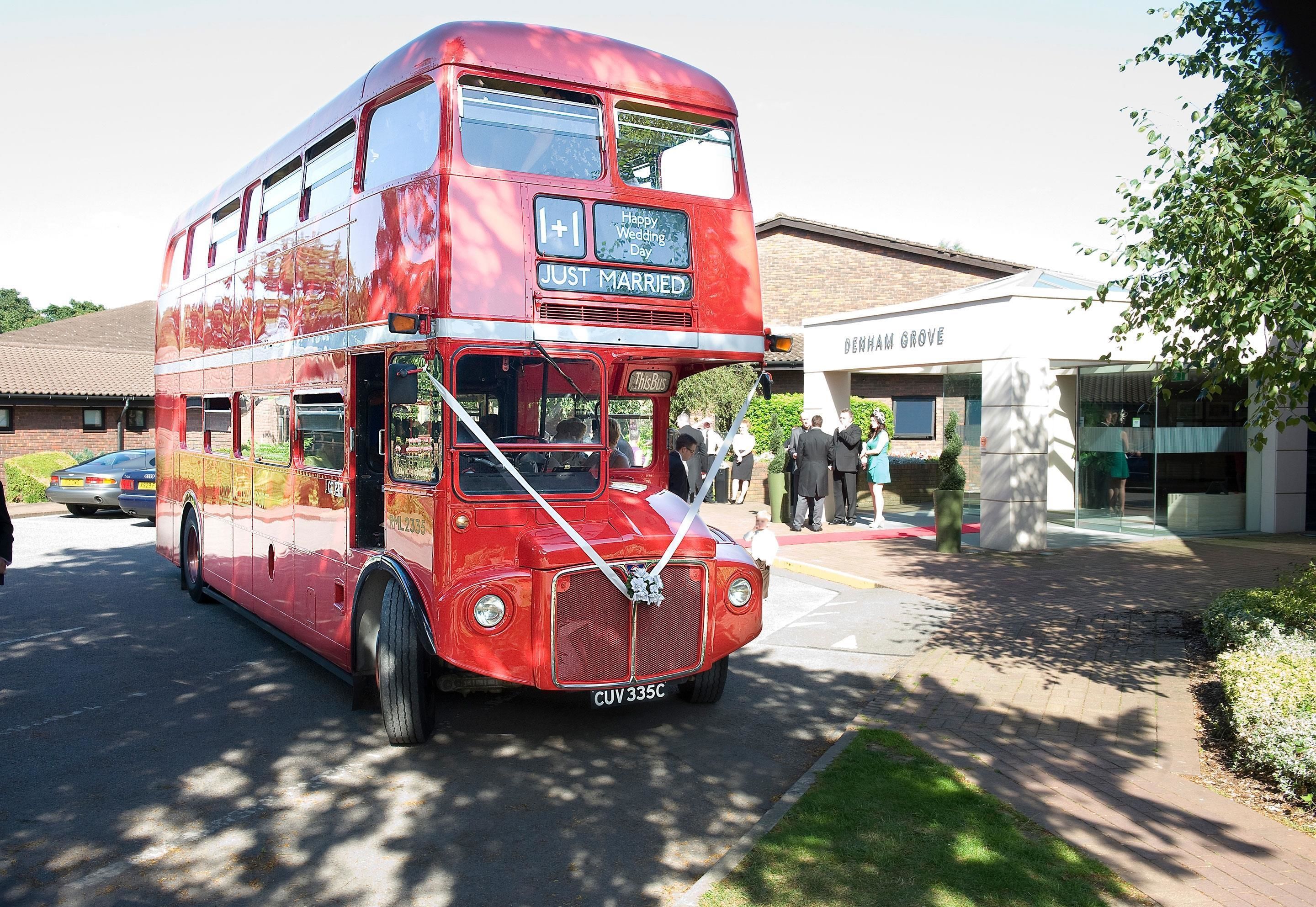 Un autobús rojo de dos pisos con letreros que dicen 'Feliz Día de Boda' y 'Recién Casados', estacionado frente a un edificio con un cartel que indica 'DENHAM GROVE'.