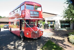 Un autobús rojo de dos pisos con letreros que dicen 'Feliz Día de Boda' y 'Recién Casados', estacionado frente a un edificio con un cartel que indica 'DENHAM GROVE'.