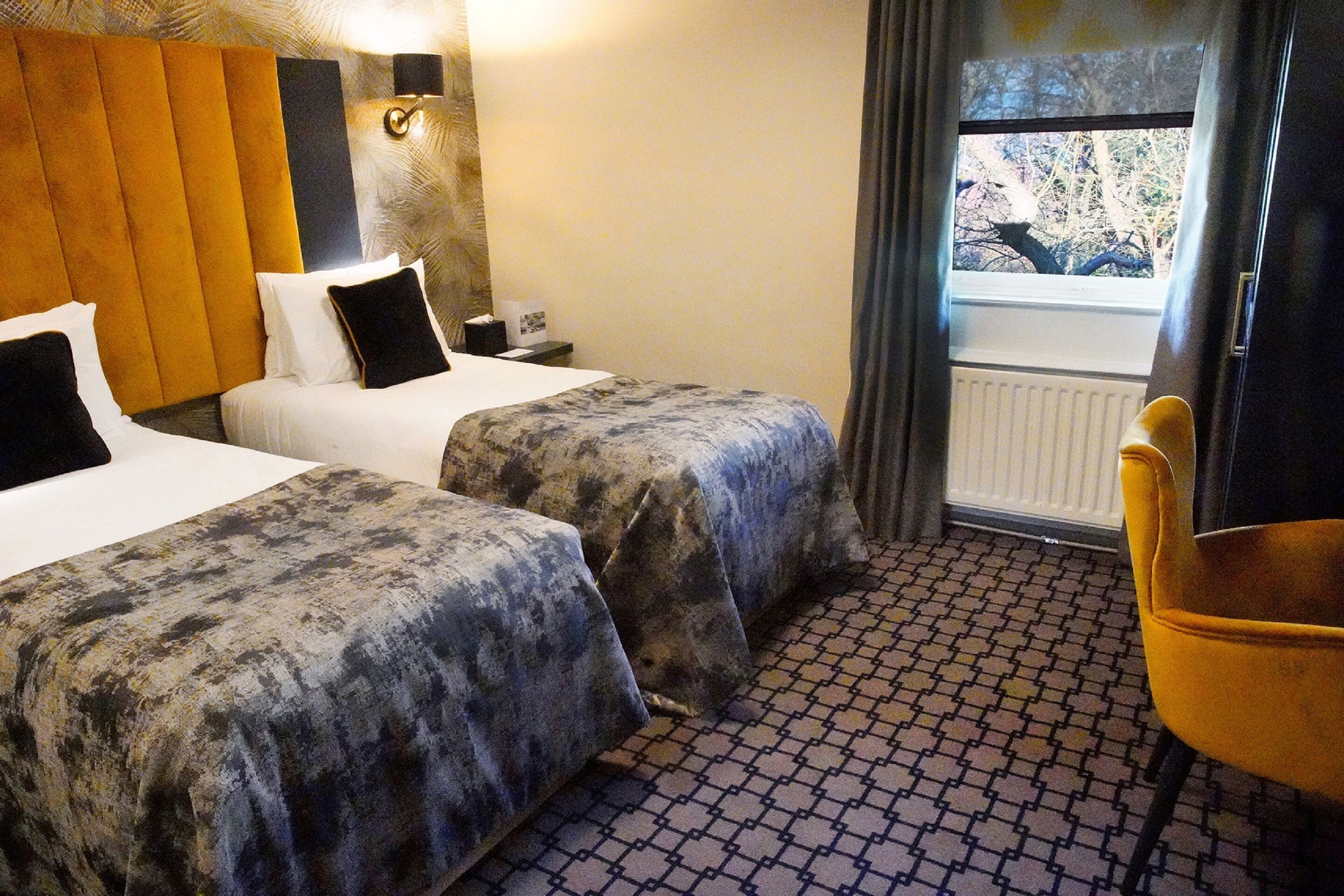 A hotel room with two beds, a small table, a chair, a window with a view of trees, and a patterned floor.