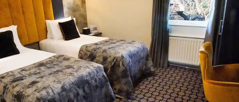 A hotel room with two beds, a small table, a chair, a window with a view of trees, and a patterned floor.