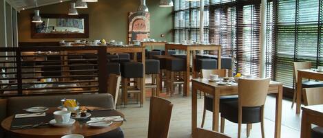 A modern dining area with multiple tables and chairs, a central bar area, and large windows with blinds.