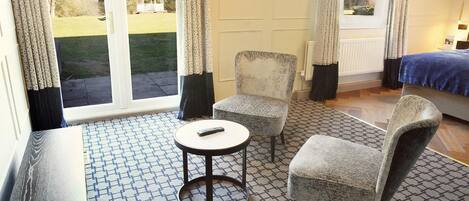 Una habitación de hotel con un ventanal grande, dos sillones, una mesita redonda y una cama con colcha azul.