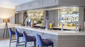 A bar area with a white countertop, bar stools, and shelves stocked with bottles and glasses. - Denham Grove (Uxbridge)