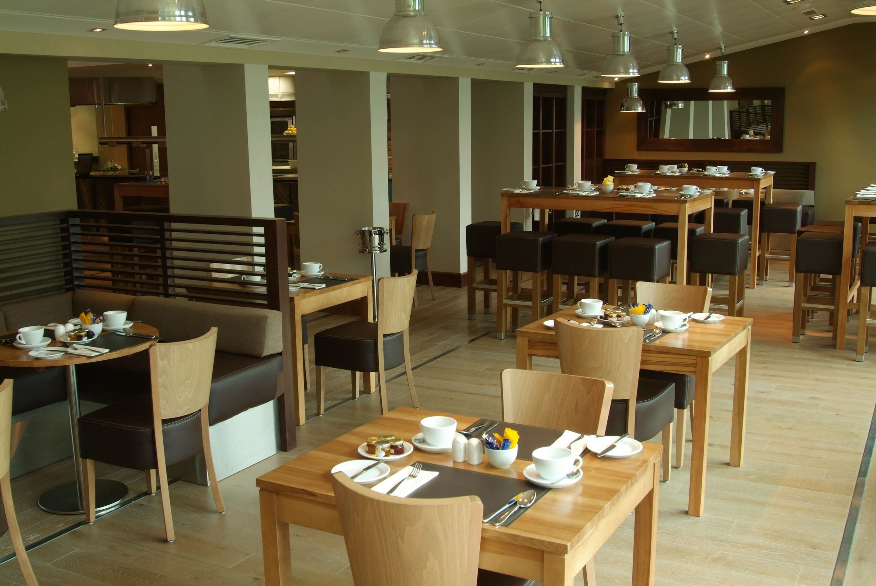 A modern dining area with wooden tables and chairs, a sofa, and a kitchen in the background.