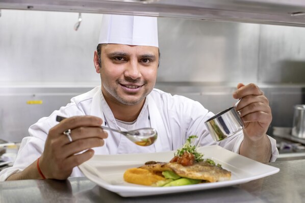 A chef in a white uniform holding a spoon and a small container, likely tasting a dish.