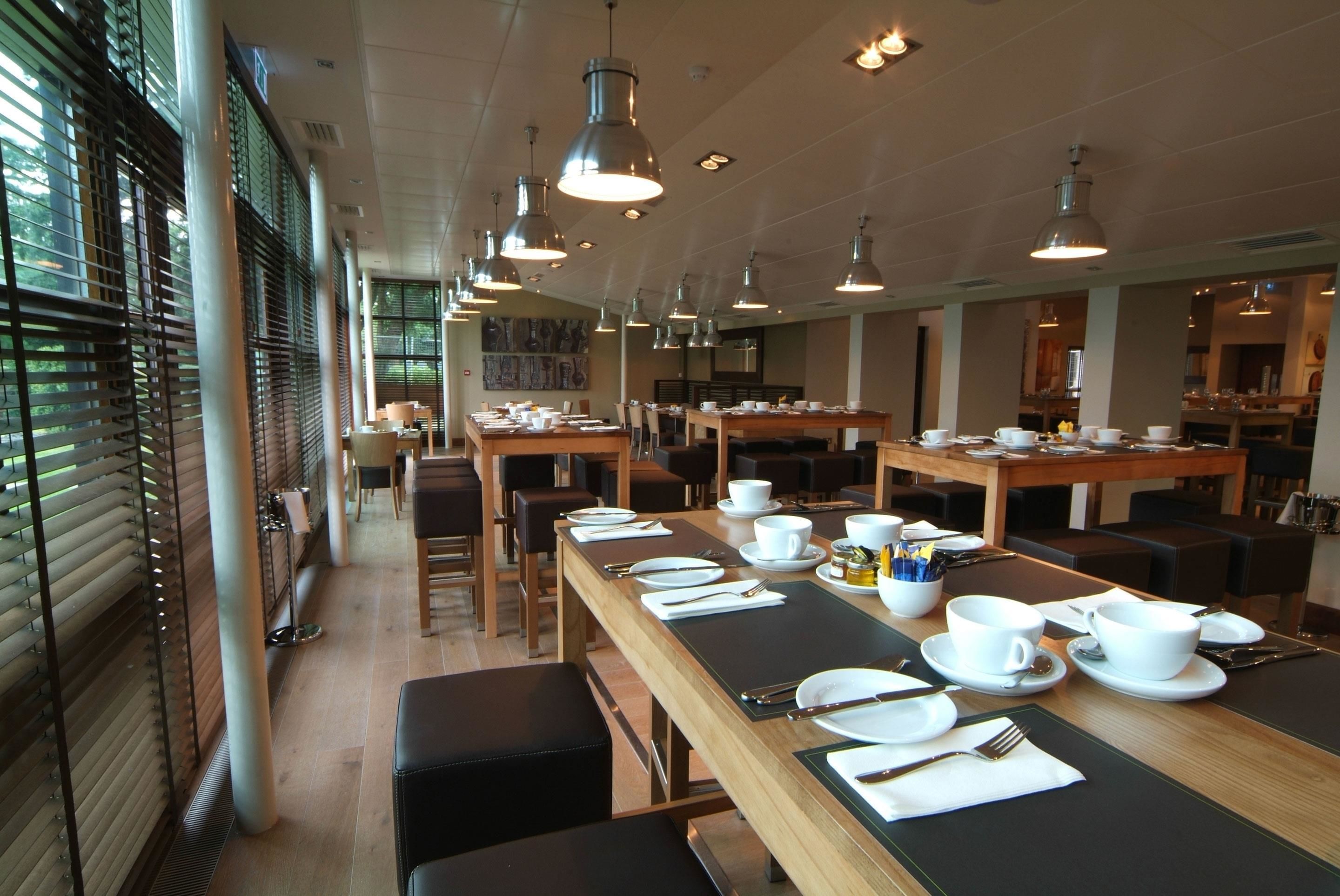 Uma ampla sala de jantar com várias mesas montadas para o café da manhã, com pratos brancos, xícaras e talheres.